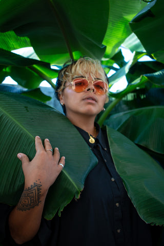 nonbinary brown person stands behind 2 huge banana leaves. they are wearing a black shirt and orange sunglasses