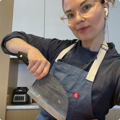 Person in kitchen holding a large knife, wearing glasses, an apron, and earbuds.