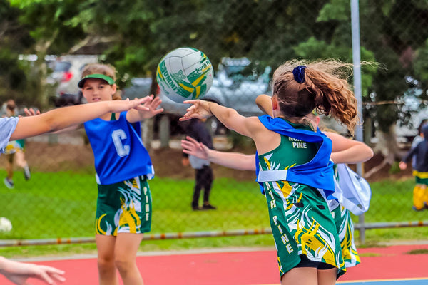 Young netbll girls passing netball in a game on court outside. Blue netball bib player passes to Centre team member.