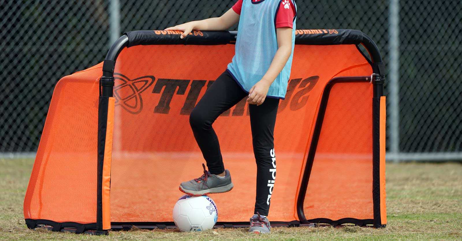 Junior soccer player standing in front of portable soccer goal by SUMMIT Sport. Young footballer has a ball under their feet and ready to play soccer using ball and portable soccer goal