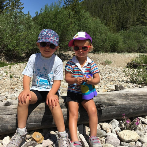 Two small kids sitting on log wearing hats and sunglasses