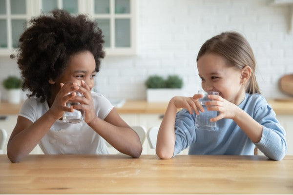 Cute smiling diverse little girls drinking fresh water, sitting at wooden table in kitchen, looking at each other