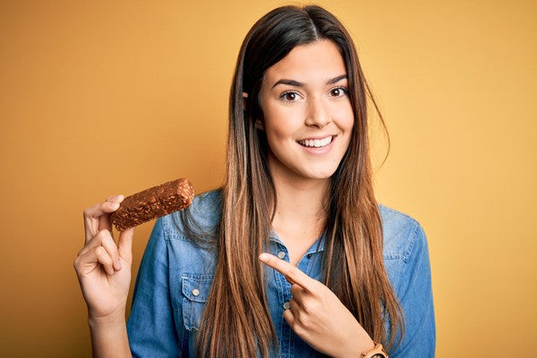 Young beautiful girl holding healthy protein bar standing over isolated yellow background very happy pointing with hand and finger