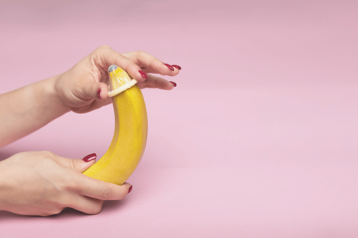 a hand sliding a condom over a banana against a light pink background