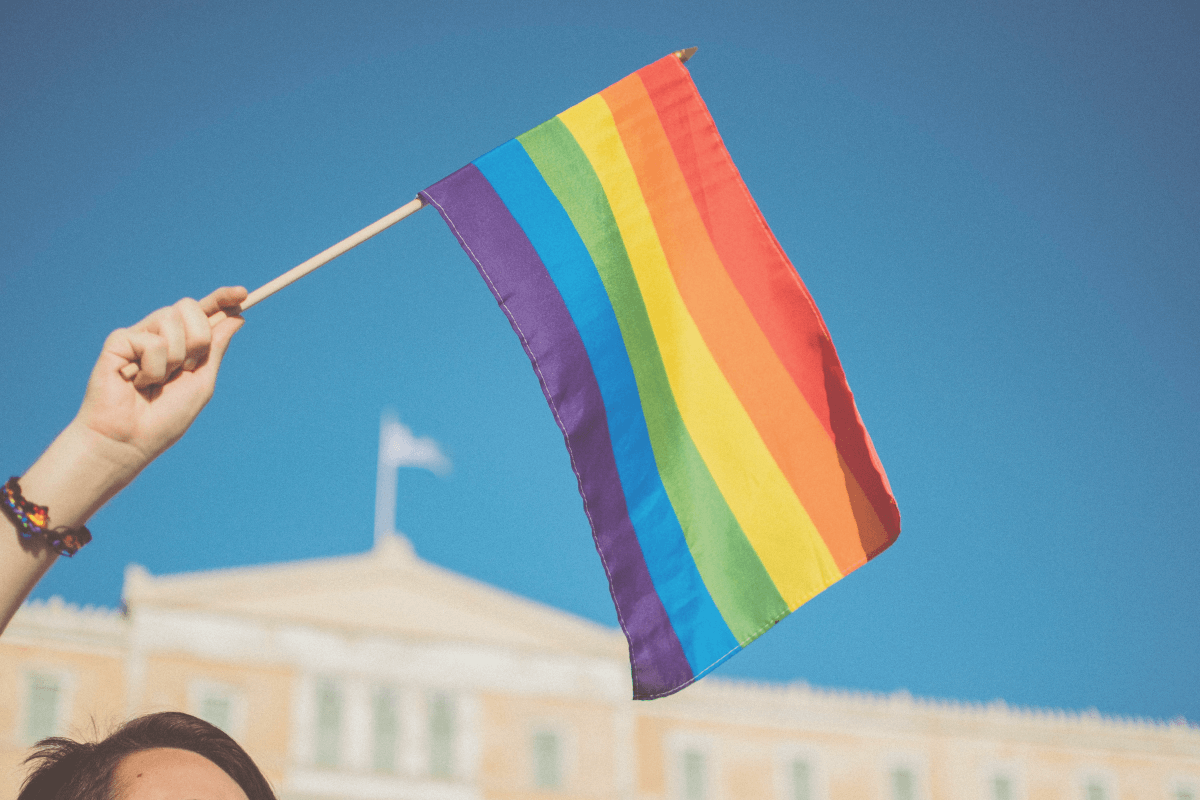 A hand holding a rainbow flag against a blue sky with a building in the background