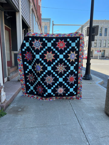 photo of a colorful quilt with a black background and turquoise diagonal chains, taken on a village street with a brilliant blue sky
