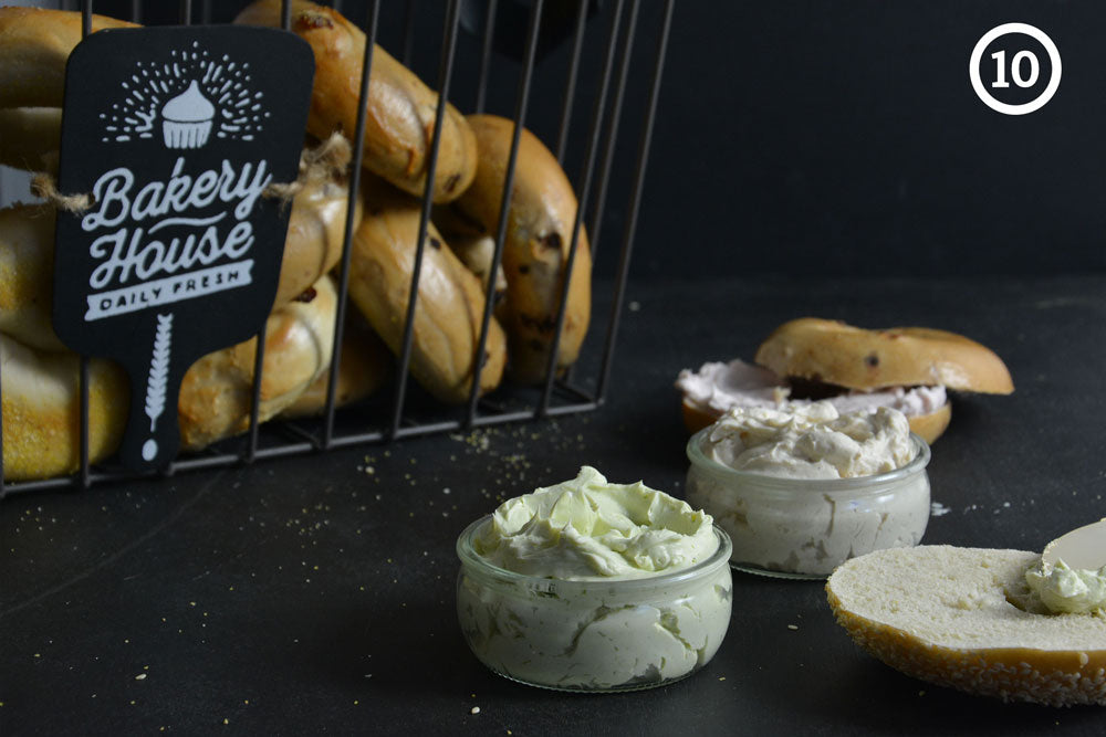 Small jars of cream cheese in the foreground with a basket of bagels in the background against a black backdrop. The image is labeled "10" in the upper right corner.