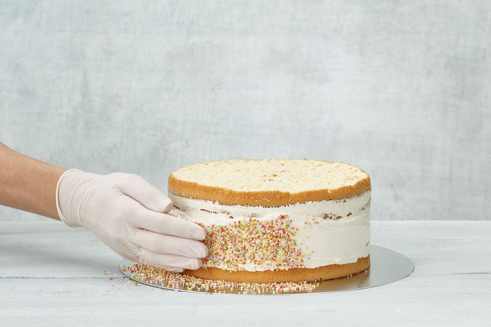 A vanilla cake with a buttercream crumb-coat being coated with multicolored sprinkles against a gray backdrop. 