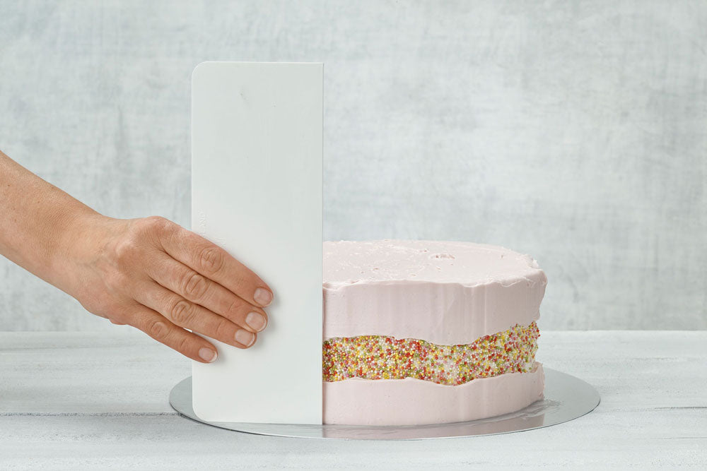 A pink cake with a multicolored sprinkle stripe across the center having it's frosting smoothed out against a gray backdrop. 