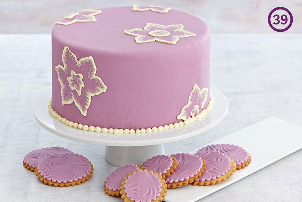 A cake decorated with purple fondant and white flowers. Cookies frosted with the same purple fondant sit below. The image is labeled "39" in the upper right corner.