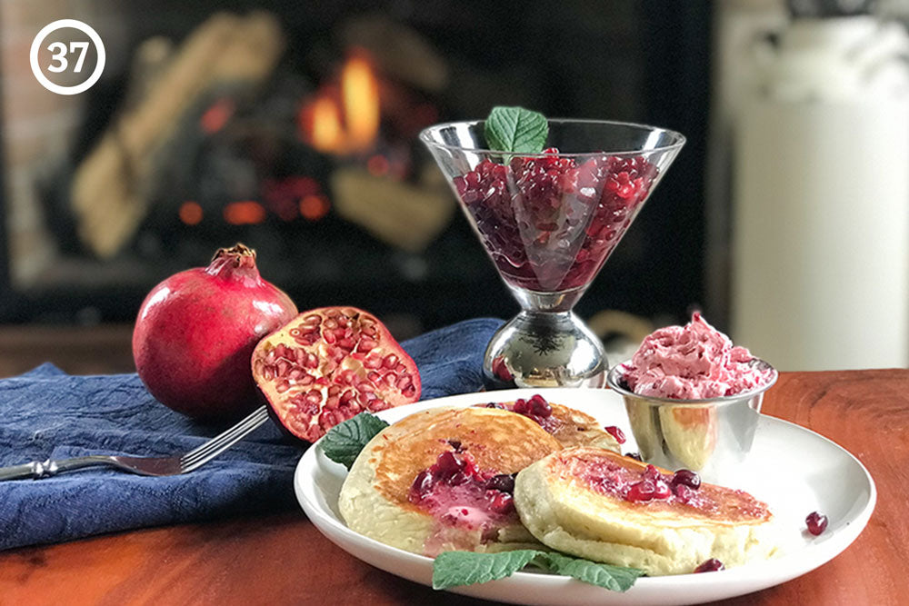 Pancakes with pomegranate butter on top in the foreground, with a whole pomegranate and a cup of pomegranate seeds in the background. A fireplace burns in the back. The image is labeled "37" in the upper left corner.