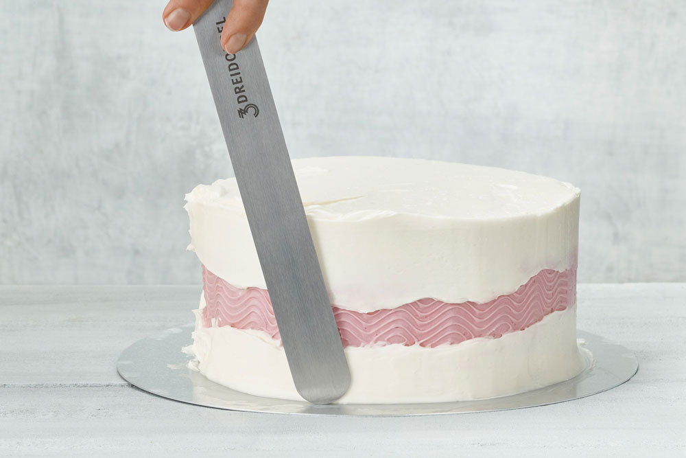 A cake being frosted with white buttercream with a wavy pink stripe across the center, against a gray backdrop. 