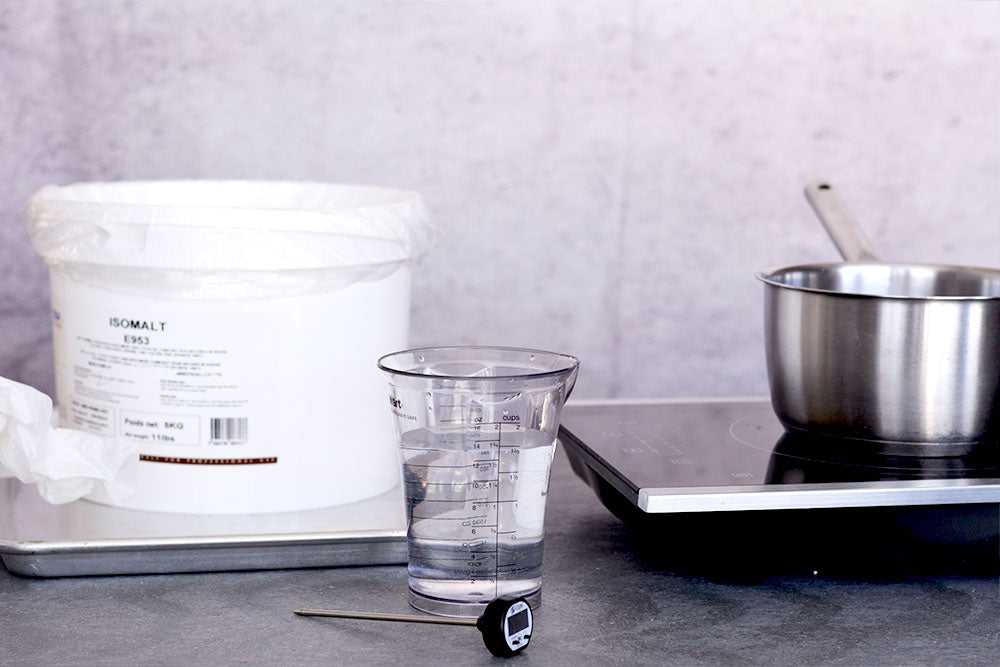 A tub of isomalt sits to the left with a pot on a stove to the right. A measuring cup of water and a thermometer sits in between. 