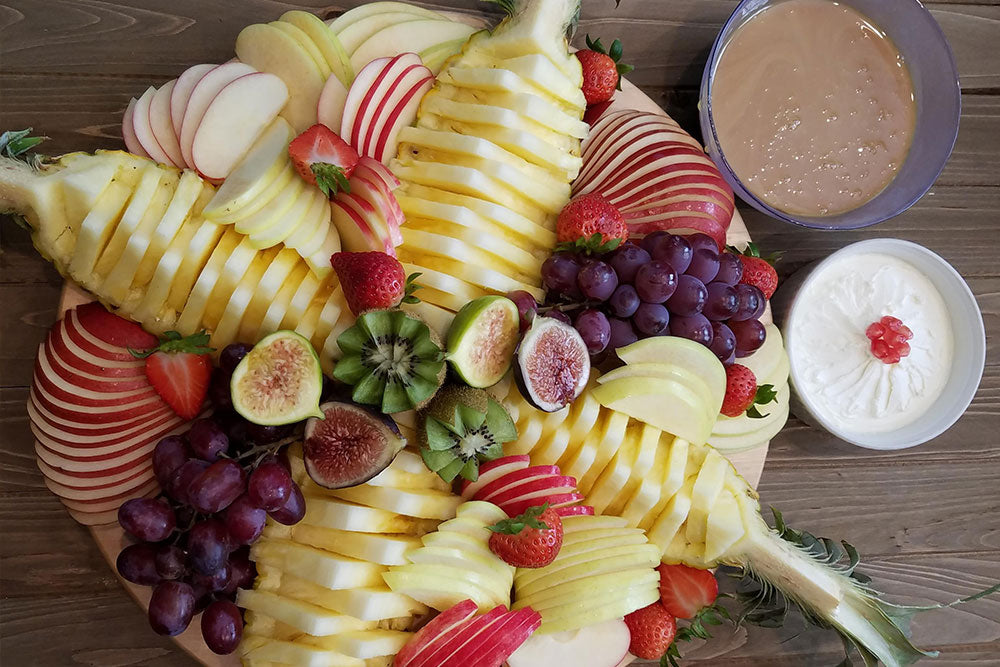 An ornate fruit plate with sliced apples, figs, kiwi, strawberries, and grapes with a bowl of pumpkin spice caramel on the side. 