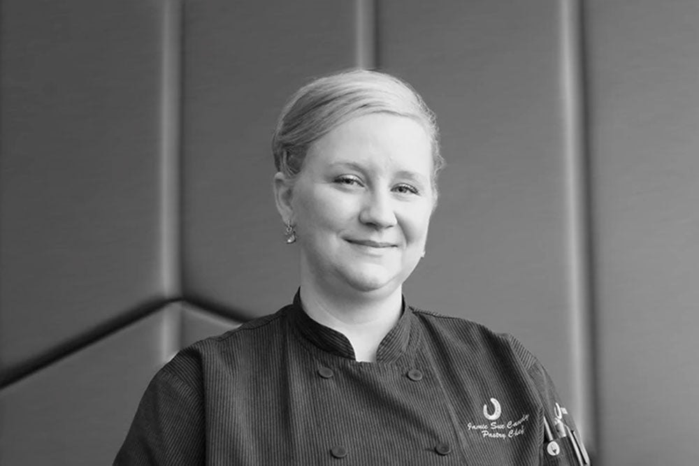 A black and white photo of a blonde woman in a dark chef's smock stands against a medium gray background. 