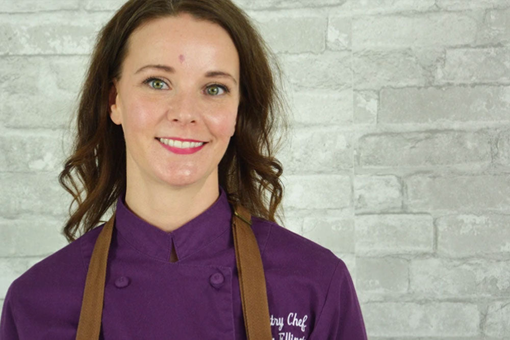 A brunette woman in a purple chefs smock and brown apron stands against a white brick backdrop. 