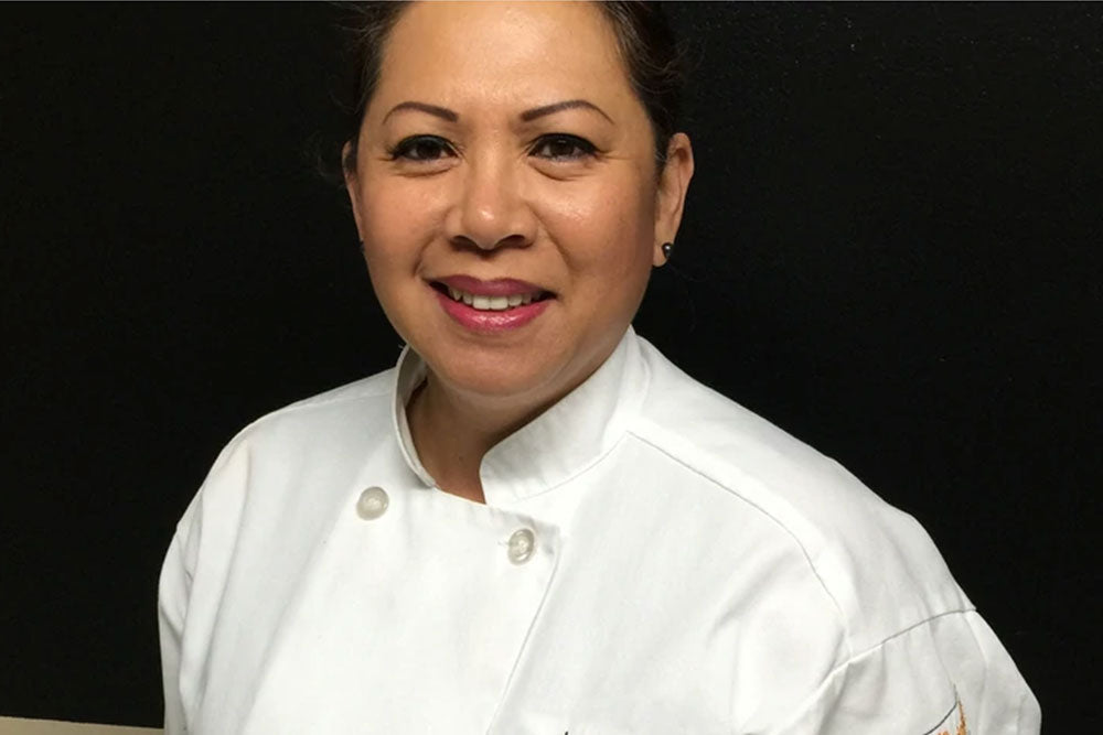 A woman in a white chef's smock stands against a black backdrop. 