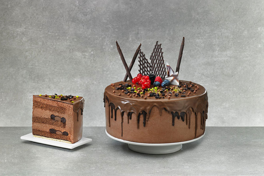 A chocolate cake with dripping ganache frosting, fresh berries a chocolate decor next to a slice of the same, against a gray backdrop. 