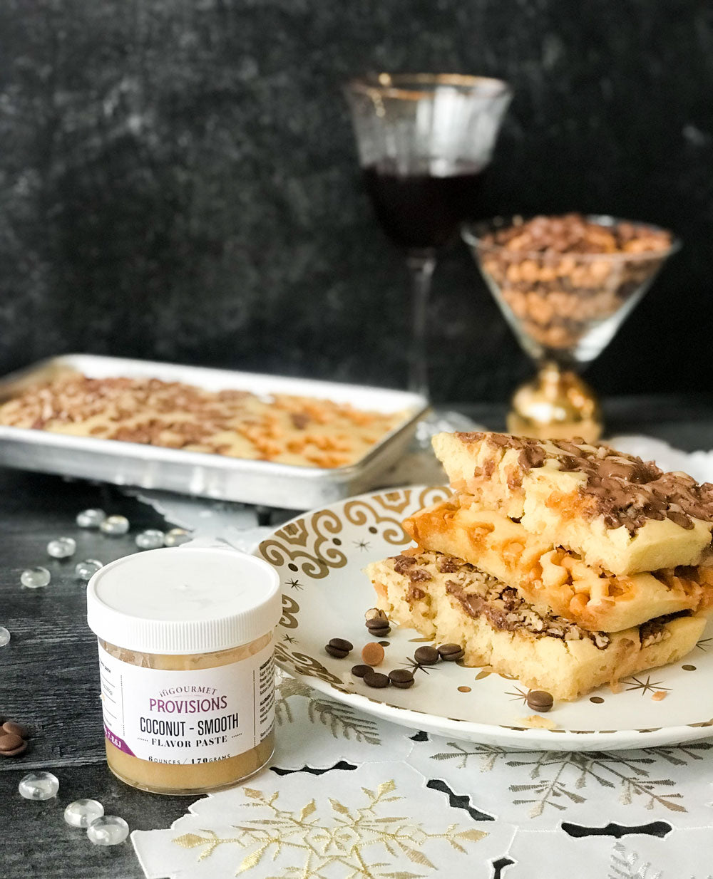 Square pancakes sit on a plate covered in chocolate chips. A jar of flavor paste sits in front and to the left of the plate. A tray of pancakes sits in the background along with a glass of wine and a bowl filled with butterscotch and chocolate chips. 