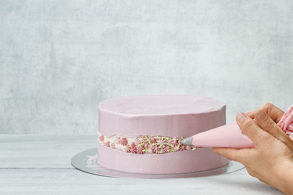 A purple cake against a gray backdrop being frosted with buttercream flowers across the center. 