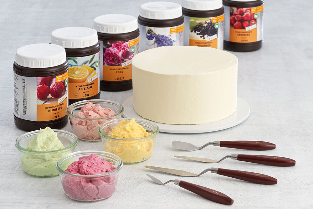 A white frosted cake backed by jars of dreidoppel flavor paste. 4 bowls of colored buttercream frosting sit next to the cake, with 4 palette knives on the other side. 