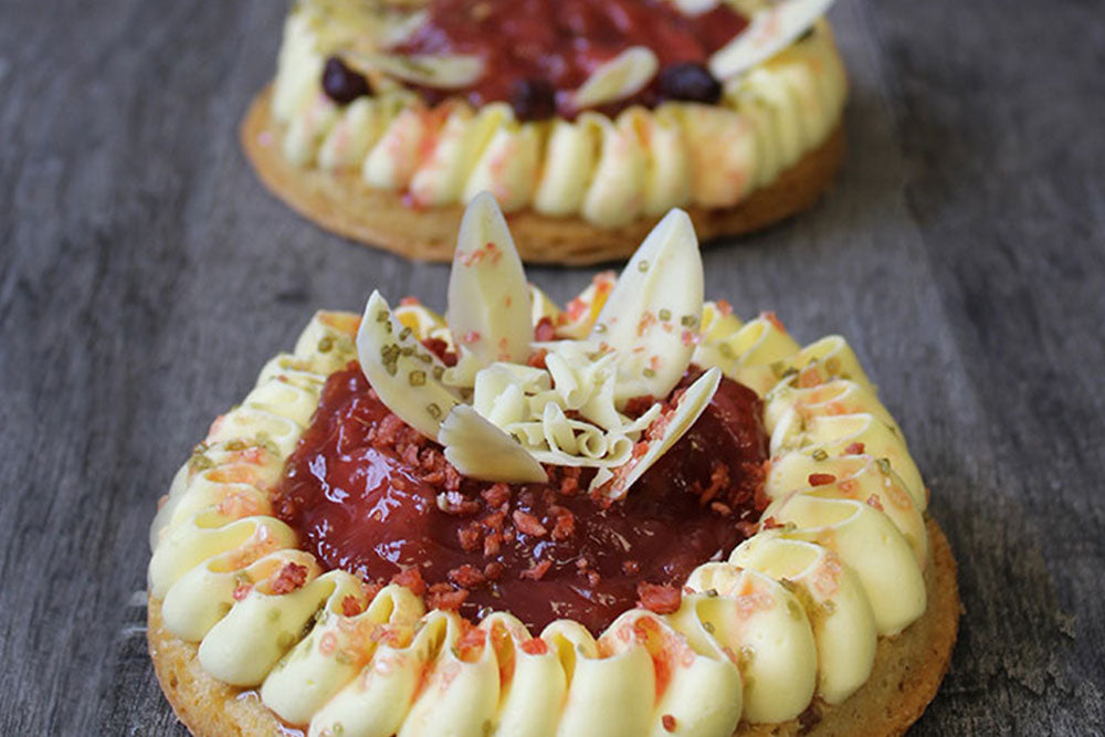 A jam tart with white ribbon icing on the side and white chocolate curls on top, sitting on a dark gray table. 