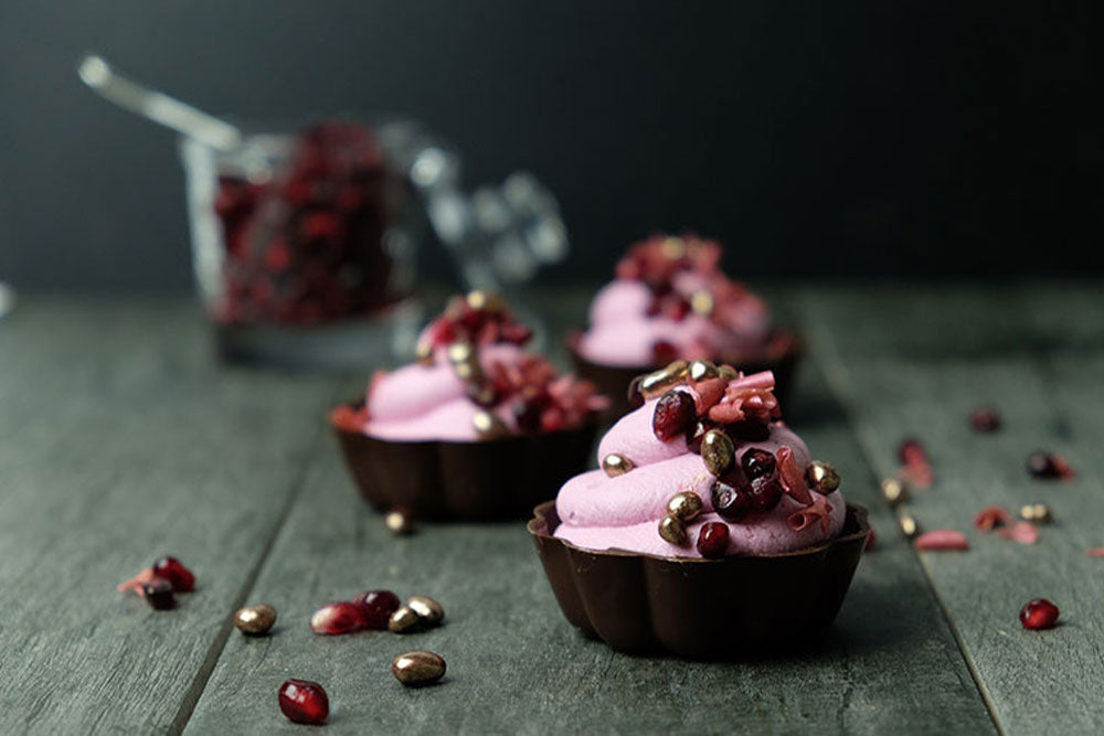 Chocolate cups filled with a pink pomegranate mousse decorates with pomegranate seeds and gold sprinkles, with a jar of pomegranate seeds in the background, all against a dark background. 