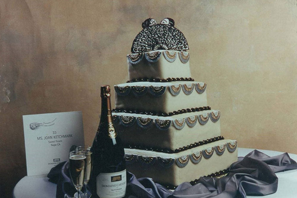 A 4 tier white wedding cake sits on a gray cloth on top of a white table. A bottle of champagne and two champagne flutes sit in front and to the left of the cake. A wedding invitation sits to the left of the cake. 
