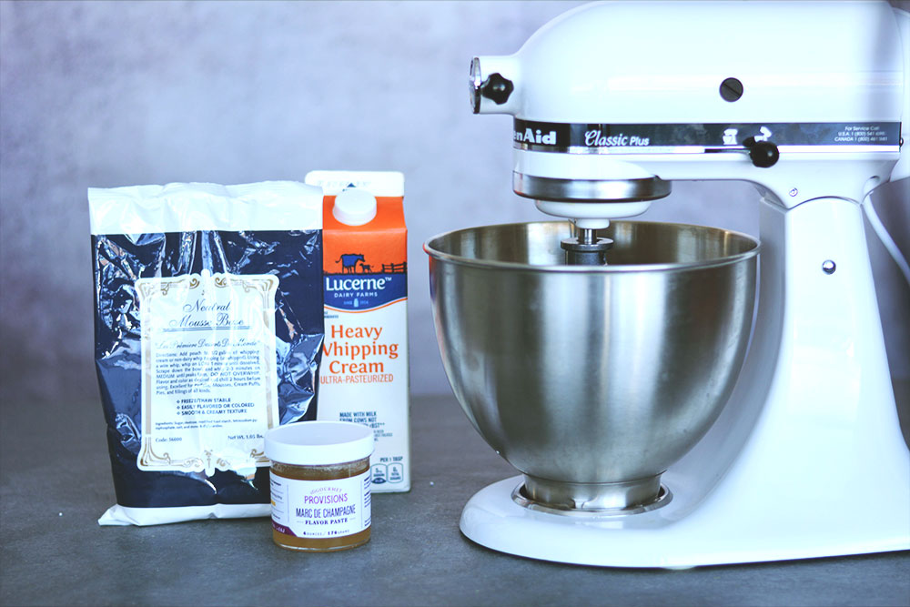 A carton of lucerne heavy whipping cream sits behind a bag of Neutral Mousse Base with a jar of flavor past in font. A White standing mixer sits to the right. 