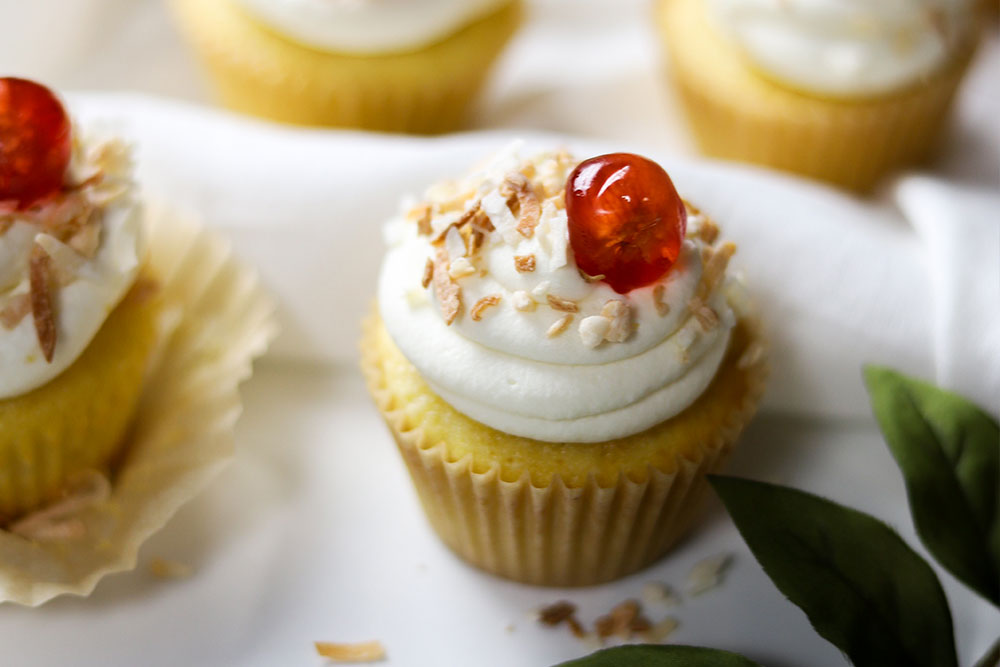 Image of soft Pineapple Coconut Cupcakes, frosted with Piña Colada Frosting and sprinkled with shredded coconut and red cherry berries.