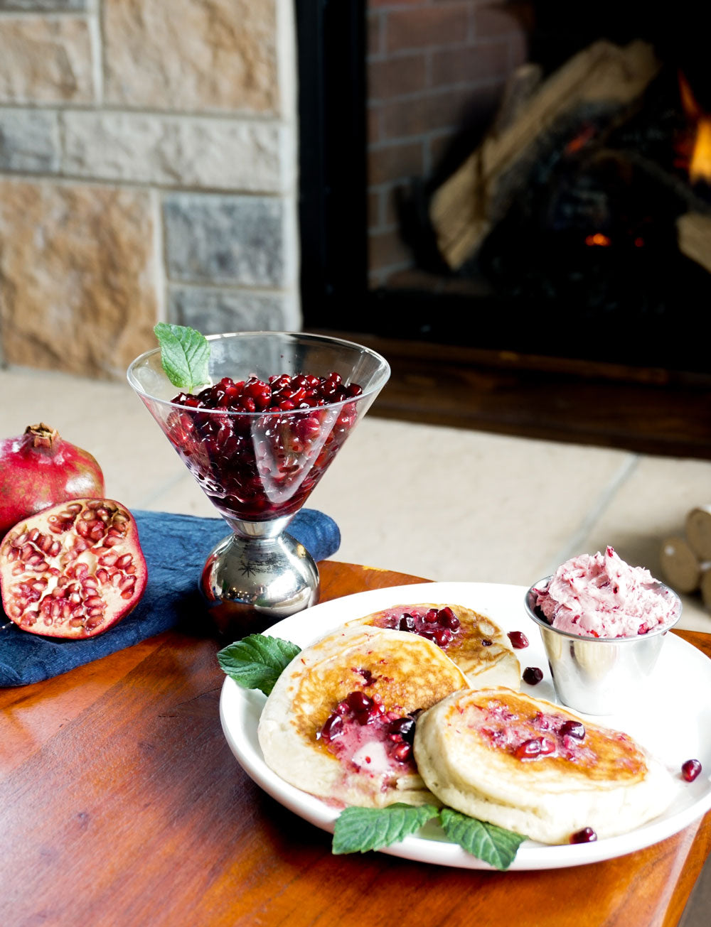 Pancakes with pomegranate butter on top in the foreground, with a whole pomegranate and a cup of pomegranate seeds in the background. A fireplace burns in the back. 