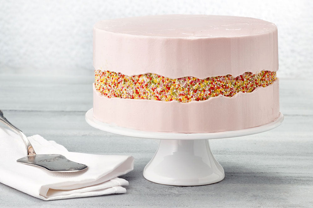 A pink cake with a multicolored stripe of sprinkles across the center against a gray backdrop. A silver spatula sits on top of a white napkin next to the cake. 