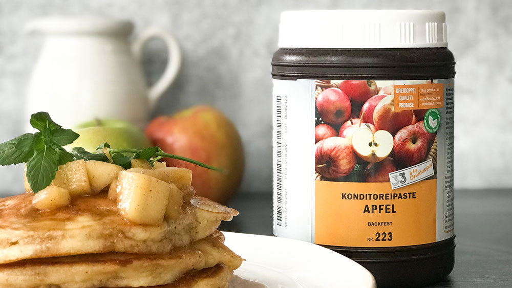 A jar of Apple flavor paste sits to the right of a plate of apple cinnamon pancakes. A white pitcher and apples sit in the background against a light gray backdrop. 