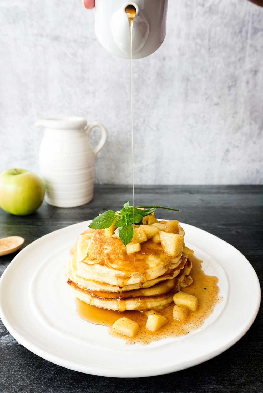 A stack of pancakes with apples on top sits on a white plate on a charcoal table. Syrup is being poured on top of the pancakes. A white pitcher and an apple sit in the background to the left against a light gray backdrop. 