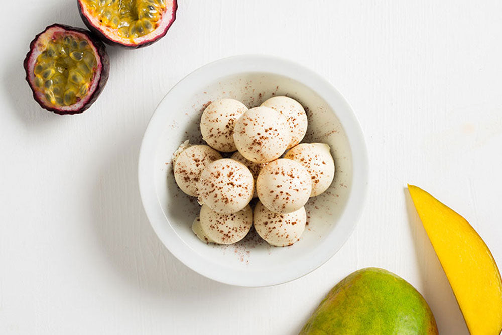 white passionfruit-mango truffles in a bowl in the center of the image, with cut open passionfruit at the top left corner of the image, and sliced mango in the bottom right corner of the image. 