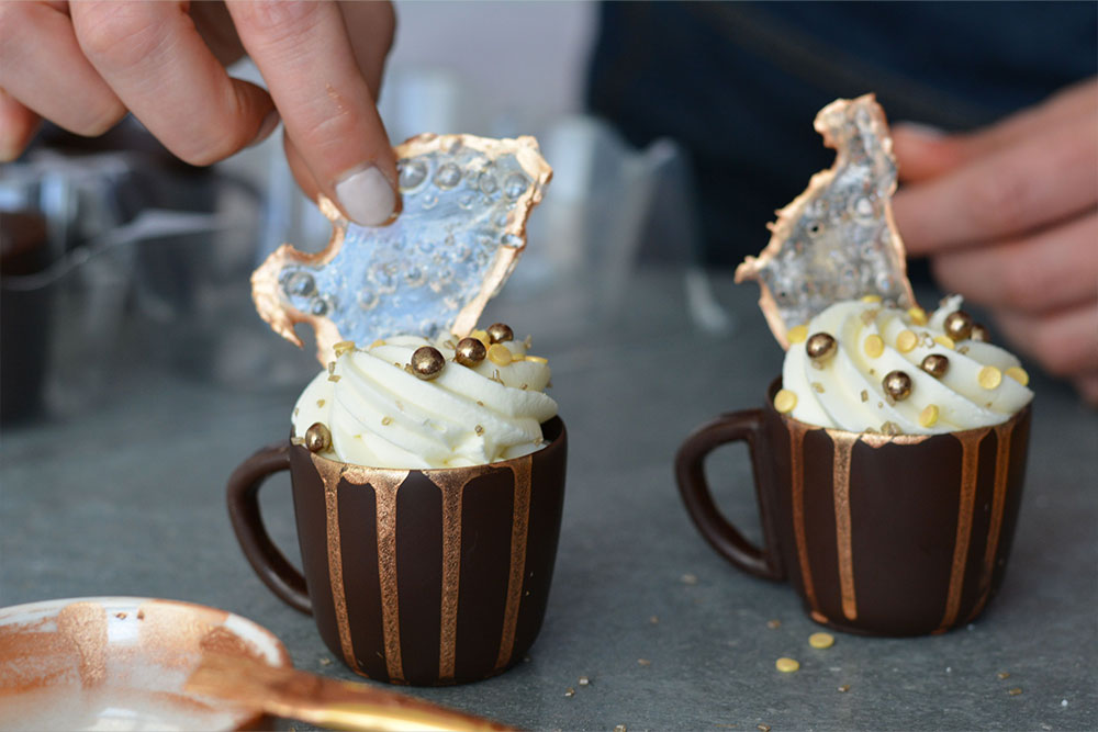 Two chocolate mugs decorated with sprinkles have caramelized sugar decorations placed on top.  