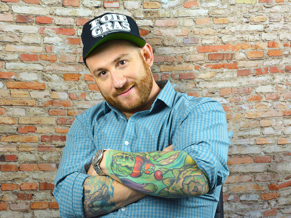 A man with his arms cross to show tattoos in a blue shirt with a hat on that says "foie gras" against a brick backdrop