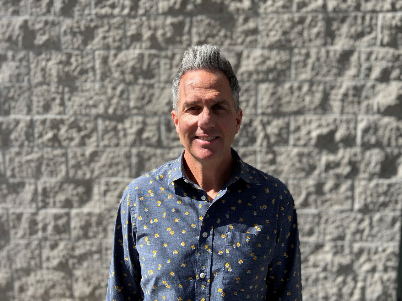 A man in a small yellow flower-patterned button down against a white brick backdrop.