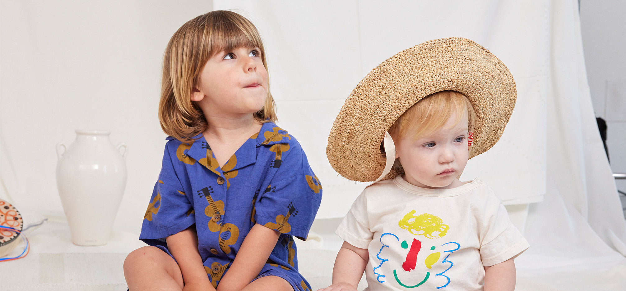 Two babies sitting by a white back drop wearing a bobo choses guitar print set and other in a straw hat and happy face t-shirt