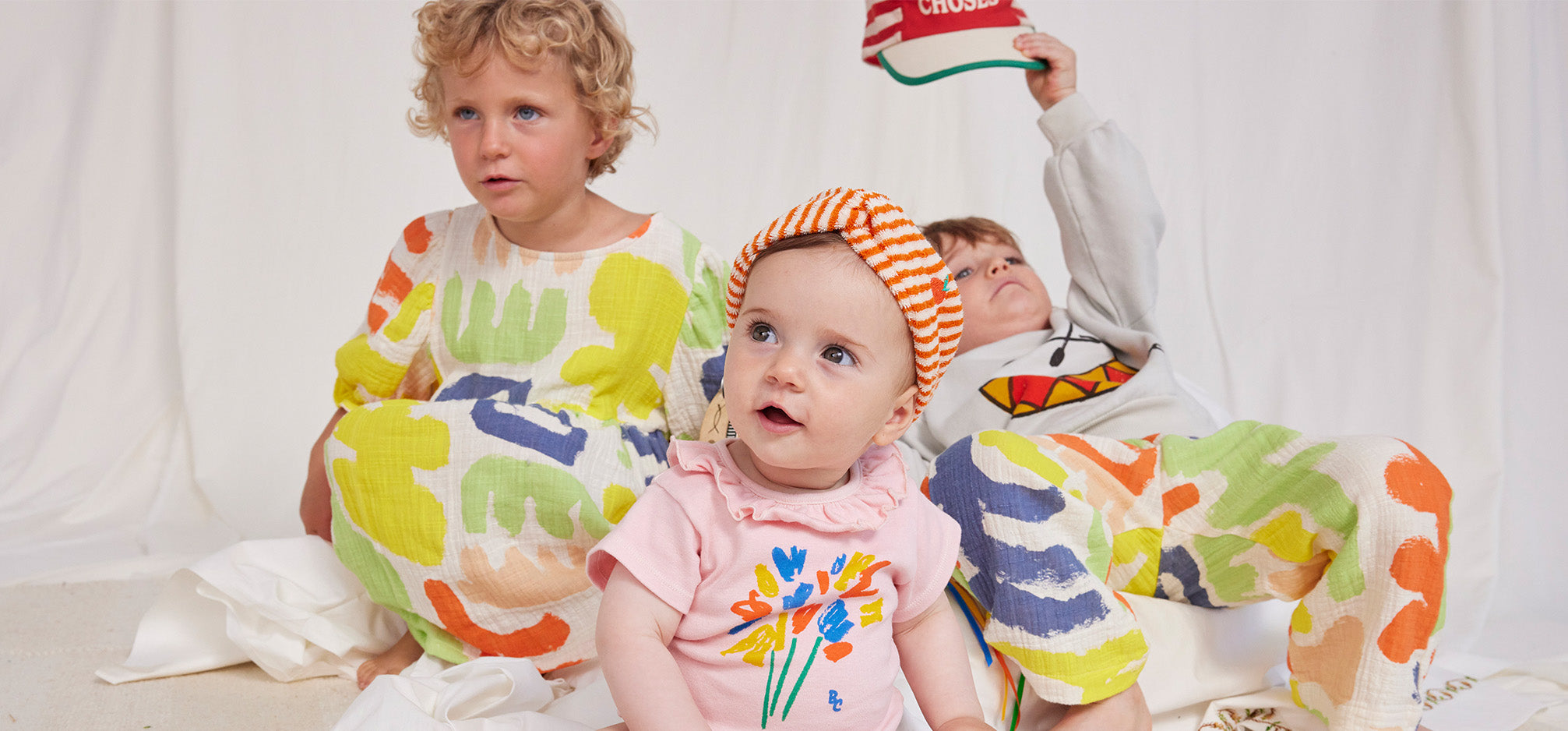 Three kids wearing bobo choses ss24 collection. Baby at the front with pink romper and striped headband.