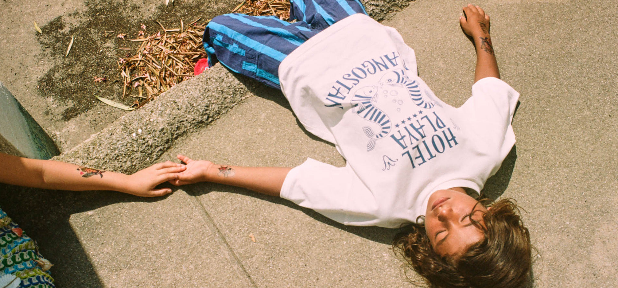 Image of child lying on concrete holding another childs hand wearing a white t-shirt with hotel playa in blue writing