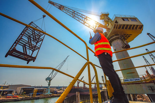 contractor with clipboard at construction site with crane in background hoisting load