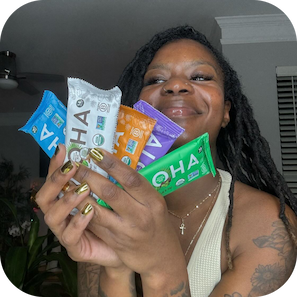 Woman smiling and holding five colorful ALOHA protein bars in a home setting.
