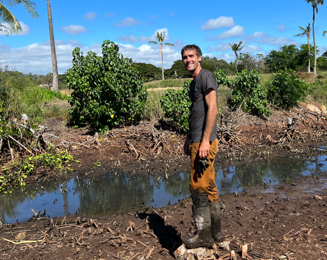 Smiling man in muddy boots standing in a field with palm trees and blue sky.