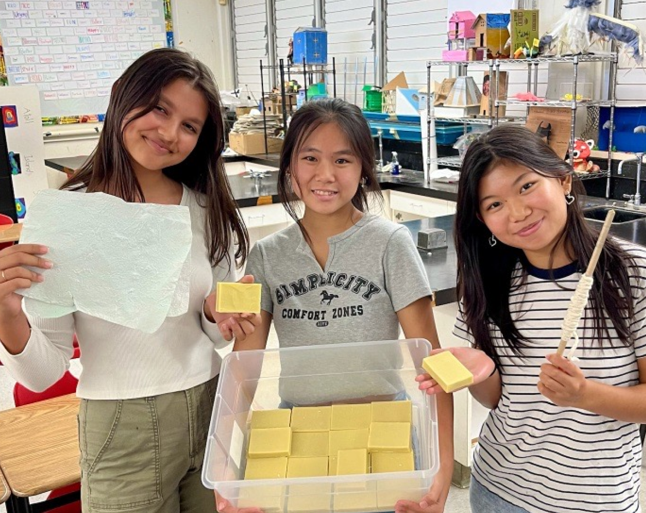 Three smiling students displaying handcrafted items in a classroom.