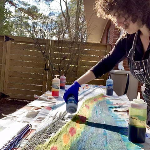 white woman applying dye in shaded of blue, green and red onto yarn with a fence in the background. 