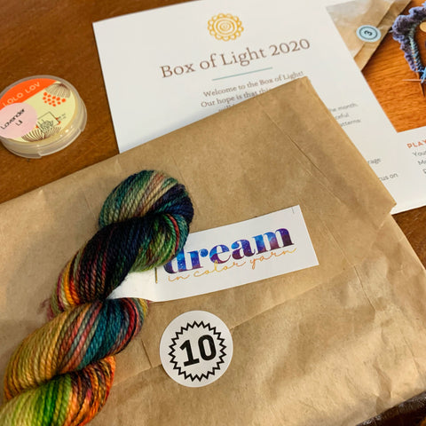 an open envelope with a number 10 sticker, a mini skein from Dream in Color, a Lolo Bar sample and a portion of the Box of Light pattern from 2020 all out on a table waiting to be worked on