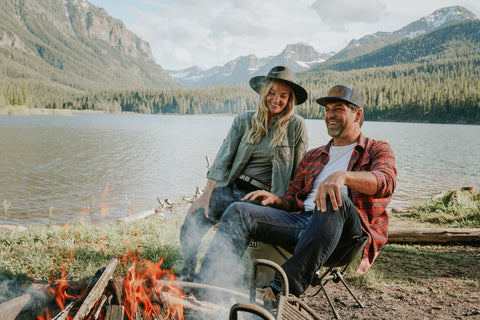 Man and woman sitting around a fire in front of mountain lake. The couple are both wearing Jelt elastic stretch belts with their mountain chic attire.