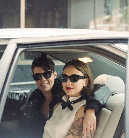 Couple in car on the way to an elopement wedding. Bride is cute wearing tuxedo style dress and black sunglasses