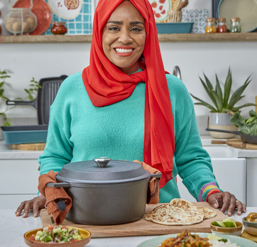 Nadiya smiles whilst standing with a cast iron casserole dish